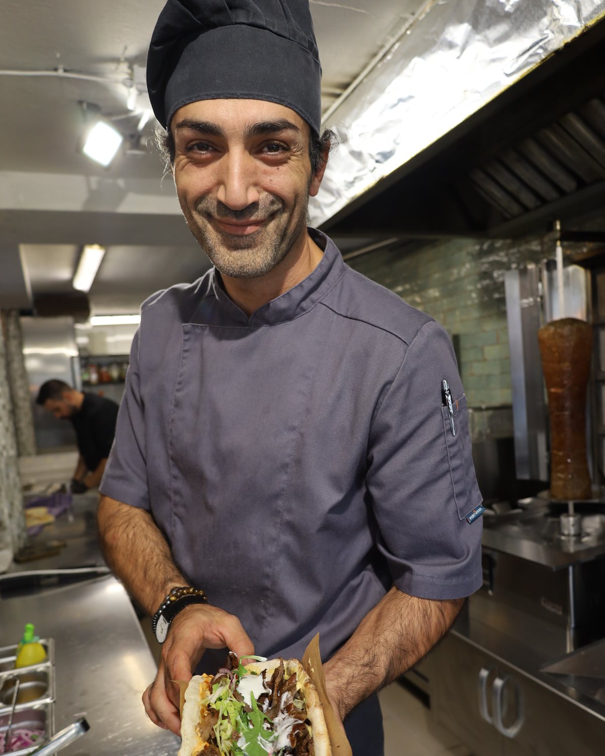 Koch von Atelier Döner hält lächelnd einen frisch belegten Steak-Döner, im Hintergrund der Spieß am Lavagrill